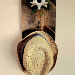 May include: A rustic wooden wall-mounted hat rack. The rack features a decorative bone and moss accent at the top, a black hook, and a straw hat with a blue band hanging from the hook. The wood has a dark stain.