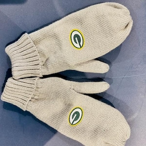 May include: A pair of beige knit mittens with a green and yellow Green Bay Packers logo on each mitt.