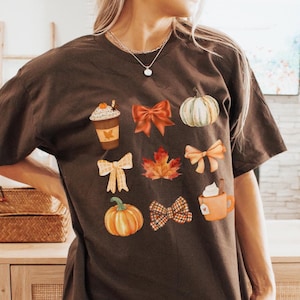 May include: A dark brown t-shirt with a fall-themed design featuring pumpkins, bows, coffee cups, and maple leaves.