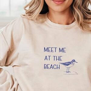 May include: A beige sweatshirt with the text "Meet me at the beach" and a small blue bird illustration.