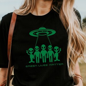 Könnte beinhalten: Ein schwarzes T-Shirt mit einer grünen Grafik eines UFOs, das über fünf Außerirdische schwebt, mit dem Text "Green Lives Matter".