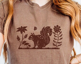 Op de afbeelding: Een bruin t-shirt met een bruine eekhoorn afbeelding omringd door bloemen en paddenstoelen.