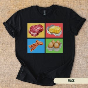 May include: A black short-sleeve t-shirt featuring a square graphic divided into four colored panels. The panels display a raw steak on yellow, a stick of butter on red, strips of bacon on blue, and a carton of four brown eggs on green.