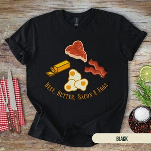 May include: A black short-sleeve t-shirt featuring a graphic of a steak, a stick of butter, bacon strips, and three fried eggs. Below the food illustrations, the text reads "BEEF, BUTTER, BACON & EGGS" in a gold-colored, arched serif font.