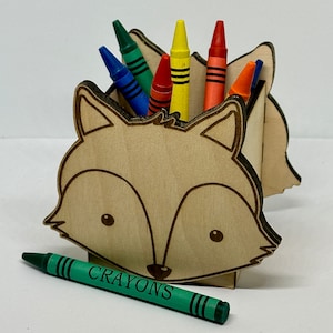 Peut inclure: Un porte-crayons en bois en forme de renard avec un crayon vert portant l'inscription "CRAYONS" devant lui. Le porte-crayons a un design découpé et est rempli de crayons de différentes couleurs, dont le rouge, le jaune, le bleu et l'orange.