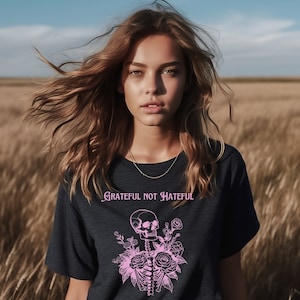 May include: A woman with long brown hair is wearing a dark blue t-shirt with a pink skull and floral design. The text "Grateful Not Hateful" is printed on the shirt.