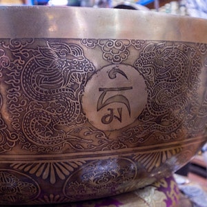 May include: A large, round, brass singing bowl with intricate engravings. The bowl features two dragons, a circular symbol, and a repeating pattern of stylized flowers. The bowl is a traditional Tibetan singing bowl used for meditation and sound healing.