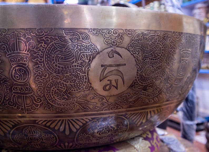 May include: A large, round, brass singing bowl with intricate engravings. The bowl features two dragons, a circular symbol, and a repeating pattern of stylized flowers. The bowl is a traditional Tibetan singing bowl used for meditation and sound healing.