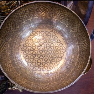 May include: A large, round, gold-colored singing bowl with intricate etched designs. The bowl has a flower of life pattern in the center and a border of repeating geometric patterns.
