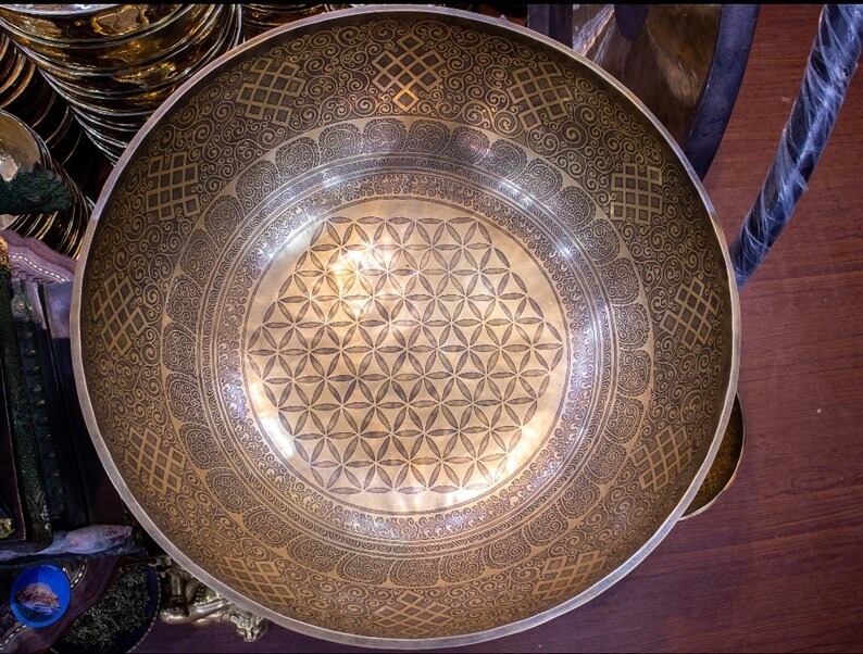 May include: A large, round, gold-colored singing bowl with intricate etched designs. The bowl has a flower of life pattern in the center and a border of repeating geometric patterns.