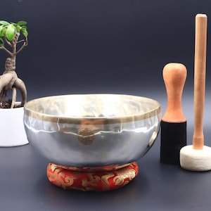 May include: A silver singing bowl with a gold rim and a gold symbol on the bottom. The bowl is sitting on a red cushion and is accompanied by two wooden strikers, one with a black felt tip and the other with a white felt tip.