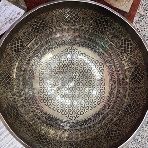 May include: A large, round, black singing bowl with a silver-colored, geometric design. The bowl has a raised, circular rim with a decorative pattern.