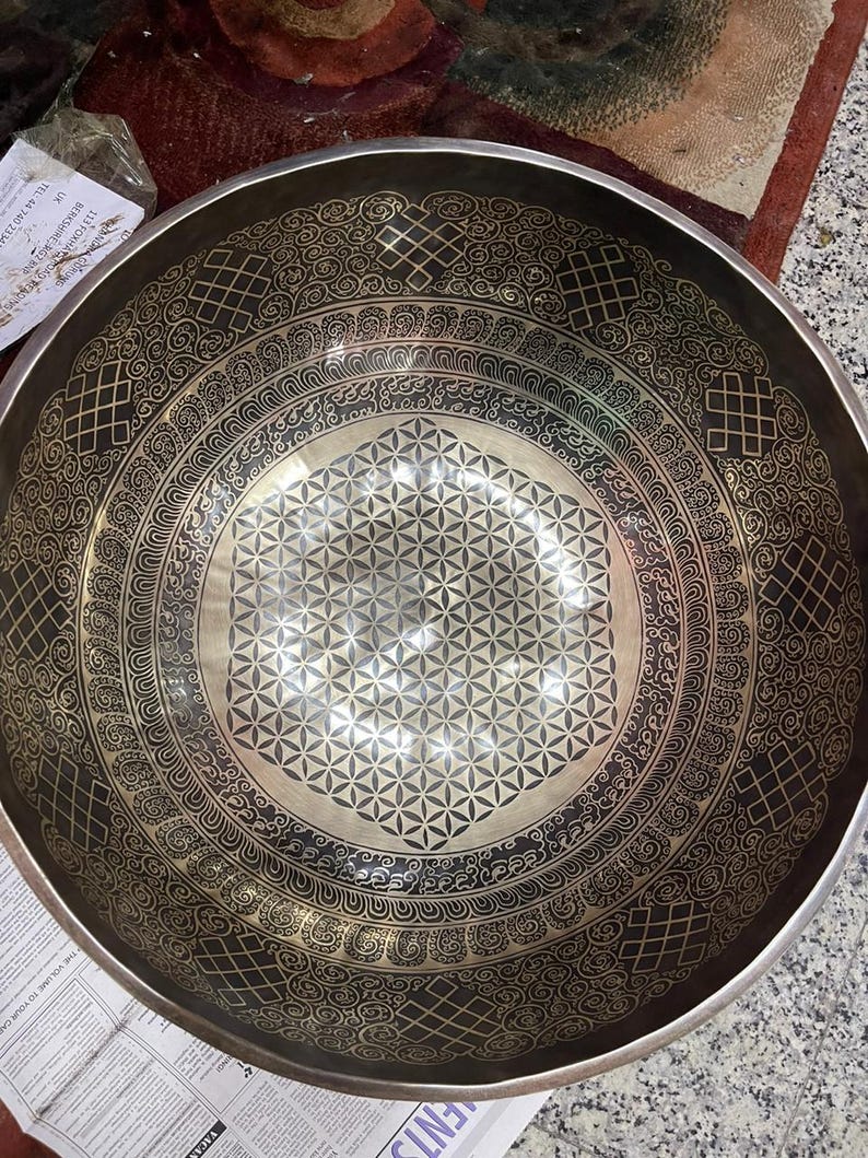 May include: A large, round, black singing bowl with a silver-colored, geometric design. The bowl has a raised, circular rim with a decorative pattern.