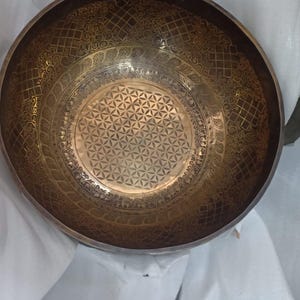 May include: A large, round, brass singing bowl with intricate etched designs. The bowl has a flower of life pattern in the center and a decorative border around the edge.