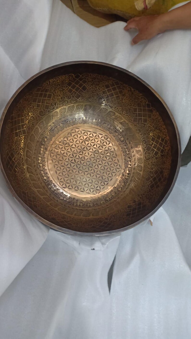 May include: A large, round, brass singing bowl with intricate etched designs. The bowl has a flower of life pattern in the center and a decorative border around the edge.