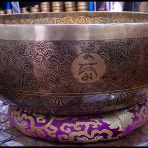 May include: A large, round, brass singing bowl with intricate engraved designs, including dragons and symbols. The bowl is resting on a purple and gold cushion.