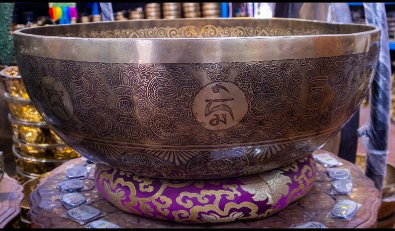 May include: A large, round, brass singing bowl with intricate engraved designs, including dragons and symbols. The bowl is resting on a purple and gold cushion.