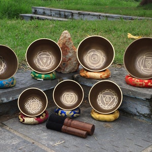 May include: Seven singing bowls with different colored cushions and a pair of wooden strikers. Each bowl has a unique design with a symbol in the center.