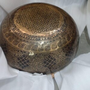 May include: A large, round, brass singing bowl with intricate designs. The bowl has a black background with gold accents and a flower of life pattern on the top.