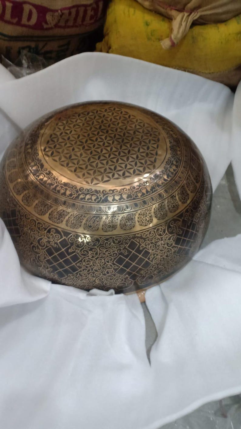 May include: A large, round, brass singing bowl with intricate designs. The bowl has a black background with gold accents and a flower of life pattern on the top.