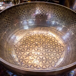 May include: A large, round, gold-colored singing bowl with intricate etched designs. The bowl has a flower of life pattern in the center and a decorative border around the rim.