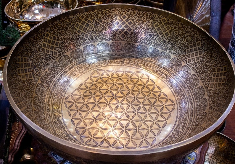 May include: A large, round, gold-colored singing bowl with intricate etched designs. The bowl has a flower of life pattern in the center and a decorative border around the rim.