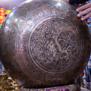 May include: A large, round, silver singing bowl with intricate engravings. The bowl features a tree of life design in the center and a border of Sanskrit characters around the edge.