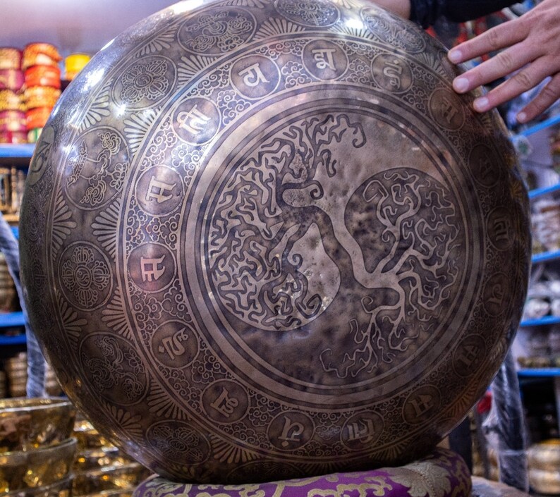 May include: A large, round, silver singing bowl with intricate engravings. The bowl features a tree of life design in the center and a border of Sanskrit characters around the edge.