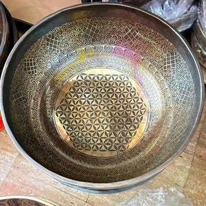 May include: A large, round, brass singing bowl with a geometric design. The bowl has a dark brown finish and a gold-colored pattern.