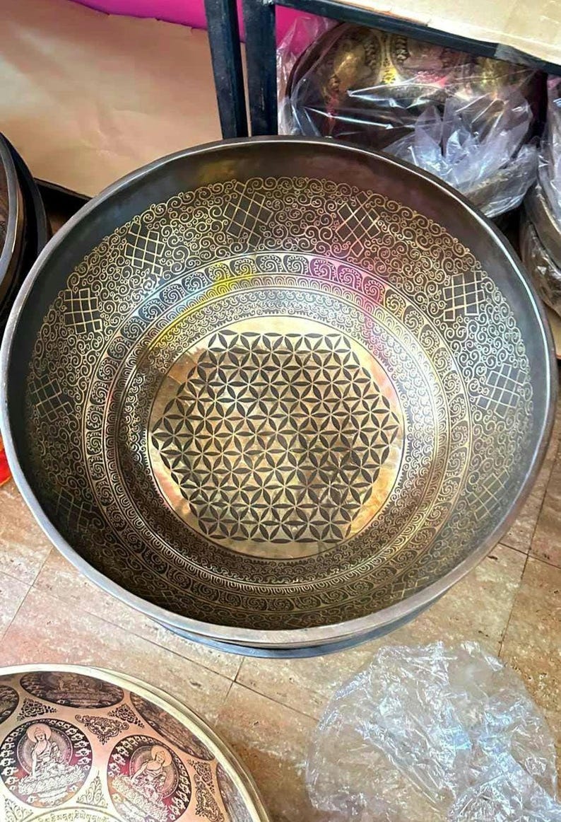 May include: A large, round, brass singing bowl with a geometric design. The bowl has a dark brown finish and a gold-colored pattern.