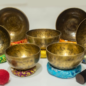 Puede incluir: Un conjunto de seis cuencos cantores de aspecto antiguo con un acabado martillado. Los cuencos están dispuestos en semicírculo sobre cojines coloridos. Se incluye un mazo de madera con una punta de fieltro roja.