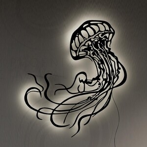 May include: A black metal wall sculpture of a jellyfish with a glowing light behind it. The sculpture is mounted on a wall with a white background.