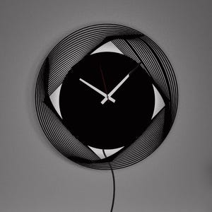 Può includere: Un orologio da parete moderno in bianco e nero con un design circolare e un quadrante nero. L'orologio è appeso a una parete grigia con una cornice bianca. C'è un vaso di rose rosse su uno scaffale sotto l'orologio.