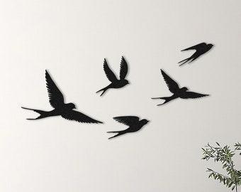 Puede incluir: Decoración de pared en silueta negra con cinco pájaros en vuelo, dispuestos en un grupo en una pared blanca. Los pájaros están todos mirando en la misma dirección, con las alas extendidas. La decoración está montada en una pared blanca, y hay un sofá y una mesa auxiliar en el fondo.
