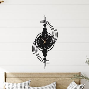Puede incluir: Reloj de pared de metal negro con una esfera circular y agujas doradas. El reloj está rodeado por un diseño decorativo de líneas curvas y círculos. El reloj está montado en una pared blanca sobre un banco de madera con cojines.