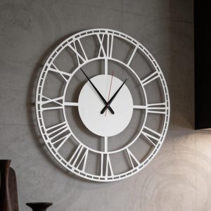 Puede incluir: Un reloj de pared blanco y redondo con números romanos. El reloj tiene un diseño minimalista con una esfera blanca y manecillas negras. El segundero es rojo. Está montado en una pared gris, creando una estética moderna y limpia. Diámetro aproximado: 40 cm.