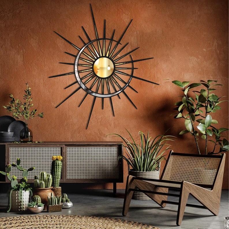 Puede incluir: Un espejo de pared de metal negro en forma de sol con un dise&ntilde;o de media luna dorada. El espejo est&aacute; colgado en una pared de terracota con una silla de madera y plantas en macetas en primer plano.