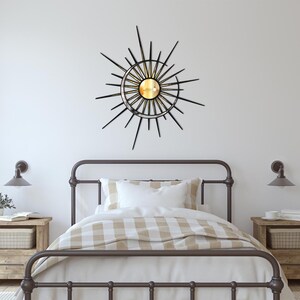 Puede incluir: Un espejo de sol de metal negro con un centro dorado y un espejo redondo en el centro. El espejo est&aacute; colgado en una pared blanca sobre un marco de cama de metal marr&oacute;n con una colcha a cuadros blancos y beige.