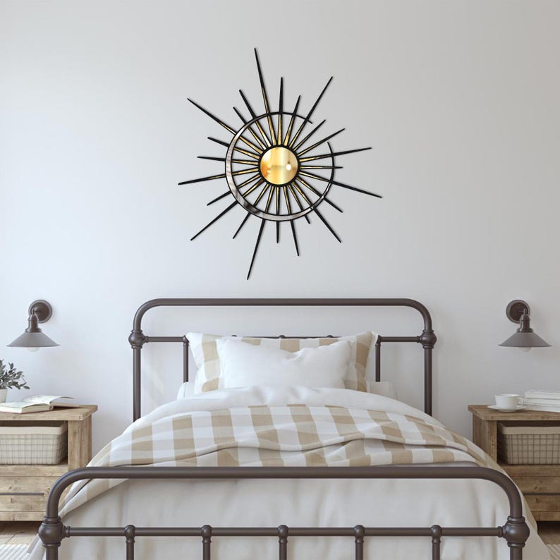 Puede incluir: Un espejo de sol de metal negro con un centro dorado y un espejo redondo en el centro. El espejo est&aacute; colgado en una pared blanca sobre un marco de cama de metal marr&oacute;n con una colcha a cuadros blancos y beige.