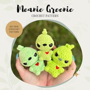 Peut inclure: Trois peluches extraterrestres vertes en crochet avec des cœurs rouges sur la poitrine. Les peluches sont tenues dans les mains d'une personne. Le texte "Meanine Greenie Crochet Pattern" est en haut de l'image.