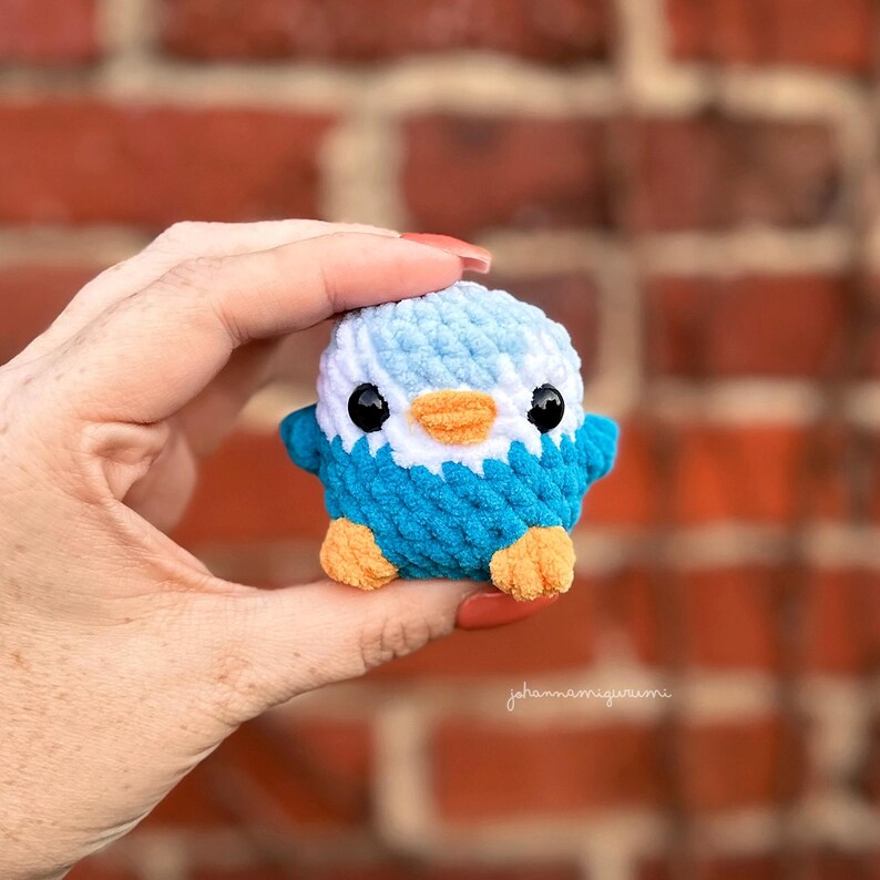 Op de afbeelding: Een klein gehaakt pingu&iuml;n speelgoed van blauw en wit garen. De pingu&iuml;n heeft zwarte ogen, een oranje snavel en oranje voeten.