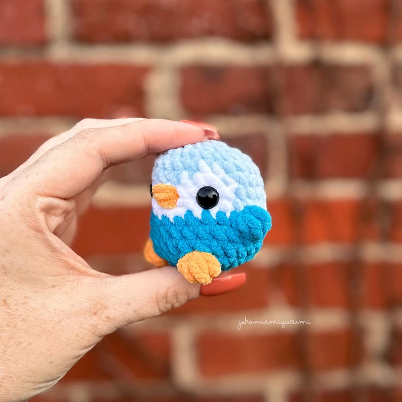 Op de afbeelding: Een klein gebreid blauw en wit vogeltje met oranje pootjes en een oranje snavel. Het vogeltje wordt in een hand vastgehouden.