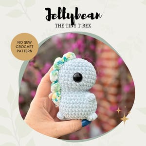 Op de afbeelding: Een lichtblauw gehaakt T-Rex-dinosaurus speelgoed met groene en gele accenten. Het speelgoed wordt in een hand gehouden. De tekst "Jellybean THE TINY T-REX" staat bovenaan de afbeelding. De tekst "NO SEW CROCHET PATTERN" staat in een cirkel linksonder in de afbeelding.