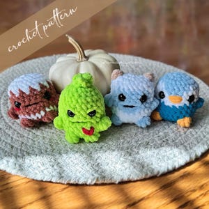 May include: Five crocheted plush toys in various colours and designs. The toys are sitting on a white crocheted doily. The toys are a brown and white gingerbread man, a green monster, a blue and white monster, and a blue penguin.