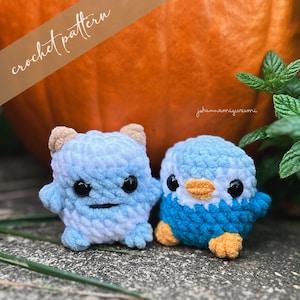 May include: Two crocheted stuffed animals, a blue and white monster and a blue penguin, are sitting on a brown surface. The monster has a white face with black eyes and a black mouth. The penguin has a white beak and orange feet. The text "jahammamigurumi" is visible in the background.