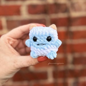 Op de afbeelding: Een klein, blauw en wit gebreid knuffeldier met zwarte ogen en een glimlachende mond. Het speelgoed heeft de vorm van een vierkant met afgeronde hoeken en twee kleine hoorns op zijn kop.