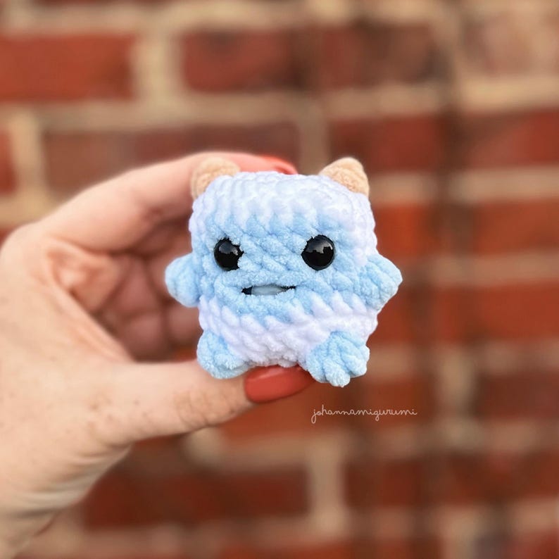 Op de afbeelding: Een klein, blauw en wit gebreid knuffeldier met zwarte ogen en een glimlachende mond. Het speelgoed heeft de vorm van een vierkant met afgeronde hoeken en twee kleine hoorns op zijn kop.