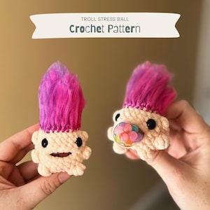 May include: Two crocheted troll stress balls with pink and purple yarn hair. One troll is holding a clear plastic ball filled with colorful beads. The image includes the text "TROLL STRESS BALL CROCHET PATTERN".