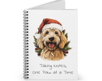 May include: A spiral bound notebook with a white cover featuring a cartoon doodle of a golden doodle dog wearing a Santa hat. The dog is surrounded by holly and berries. The text "Taking Notes, One Paw at a Time" is printed below the dog.