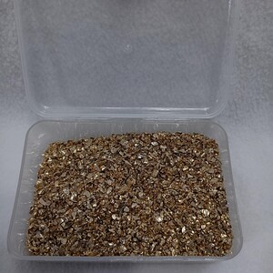 May include: A clear plastic container filled with gold glitter. The glitter is made of small, irregular pieces of gold foil.